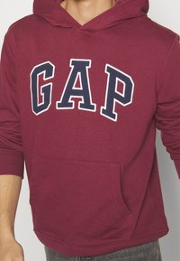 Bordowa bluza z kapturem z przednią kieszenią i dużym białym logo "GAP" obramowanym granatowym kolorem na piersi. Posiada kaptur ze sznurkiem i ściągacze na rękawach.