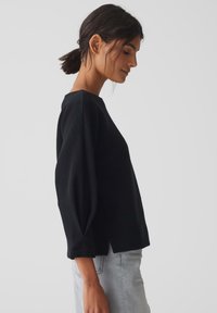 Pull en maille côtelée noire avec des manches trois-quarts, doté d'un col rond et de fentes latérales, associé à un jean gris clair.