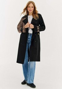 Čierny trenčkot s leopardím vzorom na rukávoch, dvojradové zapínanie. Nosený cez bielu blúzku a modré džínsy, so voľným strihom.