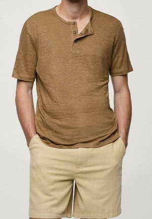 Uomo che indossa una maglietta henley marrone a maniche corte con due bottoni slacciati e pantaloncini beige, mani in tasca, in piedi contro uno sfondo semplice.