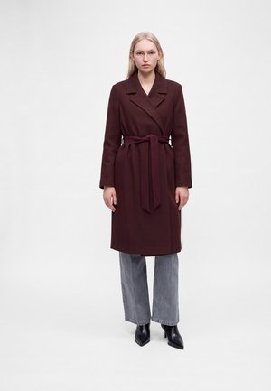 Donna che indossa un lungo cappotto bordeaux scuro con cintura, jeans grigi a gamba larga e stivali neri con tacco, in piedi davanti a uno sfondo bianco uniforme.