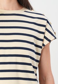 Striped topp i beige og marineblå med korte ermer, med rund hals og gullknappe-detaljer på skuldrene.