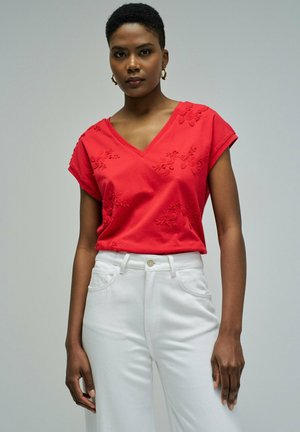 Mujer con blusa roja bordada de cuello en V y pantalones blancos de cintura alta, de pie frente a un fondo gris liso.