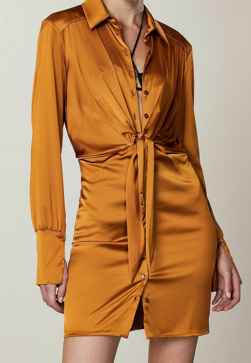 Abito a maniche lunghe, con bottoni, in satin color ambra intenso, dotato di una vita legata e di un orlo corto con bottoni discreti. Tessuto morbido e vestibilità su misura.