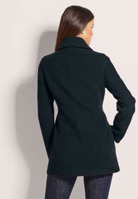 Black wool coat with a fitted design, large collar, long sleeves, and a straight back seam, extending to mid-thigh.