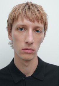 Ritratto in primo piano di una persona con capelli castano chiaro e pelle chiara, che indossa una camicia nera con colletto. Lo sfondo è bianco uniforme.