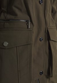 Chaqueta verde oliva de tela suave con múltiples bolsillos, cremallera frontal y botones a presión, con un acabado suave.