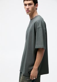 T-shirt oversize a maniche corte in tessuto grigio tenue con una texture liscia. Presenta un'etichetta del marchio all'orlo e una vestibilità rilassata.