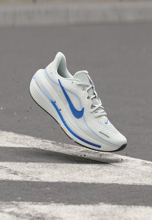 Biało-niebieski but do biegania marki Nike zawieszony nad asfaltem w pobliżu białej linii przejścia dla pieszych.