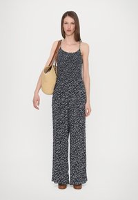 Jumpsuit floreale con sfondo nero, fiori bianchi e blu, gambe larghe e spalline sottili; abbinato a una borsa di paglia intrecciata.