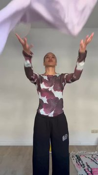 Person som har på seg en blomstret langermet topp og svarte bukser, kaster et lys lilla stoff opp i luften i et minimalistisk rom.