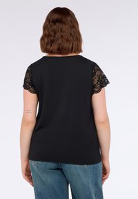 Top nero a maniche corte con delicate applicazioni in pizzo sulle maniche, texture liscia, scollatura rotonda, abbinato a jeans azzurri chiari. Visuale posteriore mostrata.