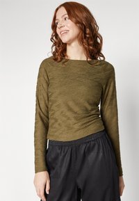 Cămașă lungă de culoare verde măsliniu cu un design texturat, ondulat. Decupaj rotund la gât și formă ajustată. Purtată cu pantaloni cu talie elastică negri.