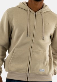Sudadera con cremallera de color beige, con capucha ajustable, dos bolsillos frontales y puños acanalados. Presenta un pequeño logotipo en el pecho y una tela texturizada.