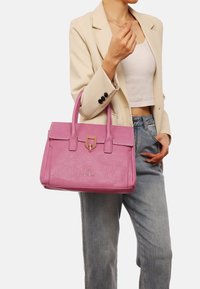 Borsa in pelle rosa con finitura testurizzata, due manici superiori, chiusura in metallo dorato e forma rettangolare. Indossata con un blazer crema e jeans.