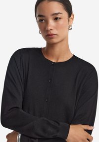 Svart cardigan i mjukt tyg, med rund hals, långa ärmar och knappstängning fram med fyra svarta knappar.