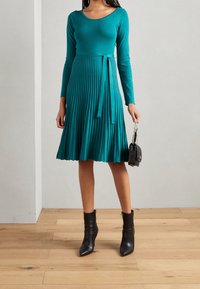 Robe midi plissée teal avec un corsage ajusté et des manches longues, cintrée à la taille par un lien assorti. Associée à des bottines noires.
