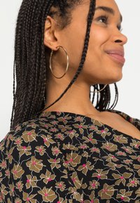Blusa preta com padrão floral que apresenta detalhes em dourado e rosa, design com decote em V, tecido suave, e cabelo trançado com grandes brincos de argola.