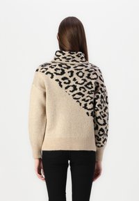 Pull beige à col montant, avec un motif léopard noir sur un côté et un tissu en tricot texturé. Ourlet et poignets côtelés.