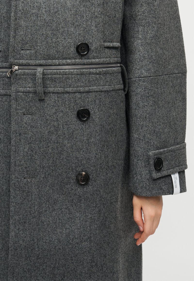 Manteau en laine grise à finition texturée, doté de boutons noirs double boutonnage, d'une fermeture éclair à l'avant et d'une taille cintrée.