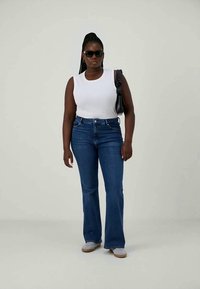 Žena v slnečných okuliaroch, bielom bezrukávovom topu, modrých džínoch, bielych teniskách a nesúca čiernu ramennú tašku na neutrálnym pozadí.