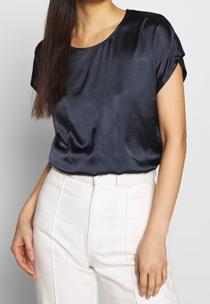 Femme portant un chemisier court en satin noir rentré dans un pantalon blanc taille haute, debout avec un bras relâché.