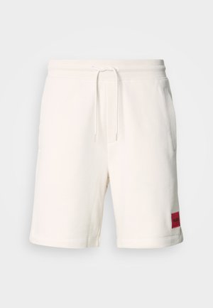 Shorts de algodón color crema con una cintura elástica, cordón de ajuste y bolsillos laterales. Presenta un parche de marca rojo en el muslo izquierdo.