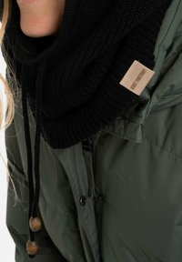 Zwarte gebreide kraagwarmer met een textuurpatroon, voorzien van verstelbare trekkoorden en houten accenten, gedragen over een groene gewatteerde jas.