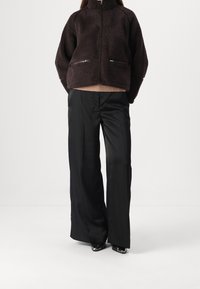 Giacca in pile marrone con colletto alto e tasche con doppia zip, abbinata a pantaloni neri a gamba larga e scarpe eleganti. Tessuto testurizzato e vestibilità rilassata.
