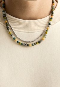 Dwie naszyjniki nałożone na siebie: jeden z wielokolorowymi koralikami (zielonymi, żółtymi, szarymi), a drugi to srebrny łańcuszek, noszone na beżowym swetrze.