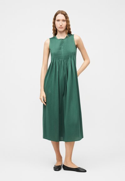 Γυναίκα με κυματιστά μαλλιά που φοράει πράσινο midi φόρεμα χωρίς μανίκια με πιέτες και μαύρα επίπεδα παπούτσια, στέκεται μπροστά σε ένα απλό φόντο.