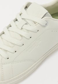 Balti odiniai sportbačiai su plokščiomis raišteliais, tekstūruotomis detalėmis ir siuvinėtu raštu. Yra logotipas, įspaustas šone, ir minimalus perforacijų kiekis.