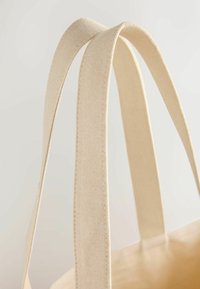 Gros plan sur deux anses de sacs fourre-tout en tissu beige sur fond neutre, montrant les détails de la texture et des coutures.