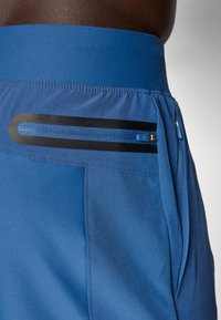 Blå idrettsshorts med en jevn tekstur, med en glidelås lomme med svarte detaljer og en kontrasterende blå glidelås.