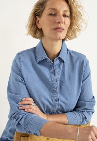 Helles blauen Denimhemd mit Button-Down-Kragen, langen Ärmeln und Ein-Knopf-Manschetten. Das Modell hat welliges Haar und ruht die Arme auf einem Holzstuhl.