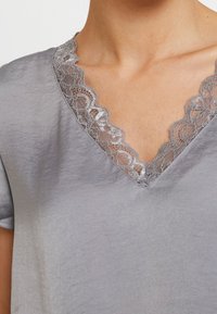 Top de satén gris con escote en V, adornado con un borde de encaje plateado. La tela tiene una textura suave con un ligero brillo. Incluye mangas cortas.