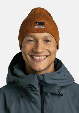 Knitted orange beanie with a mountain logo patch, worn with a gray jacket. The beanie has a textured ribbed design and a pointed top.
