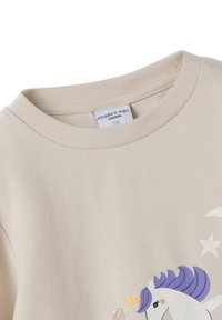 Beige kinderhemd met een eenhoorn met paarse manen en een maansterafbeelding, ronde halslijn, en maat 116 label van Polarn O. Pyret Zweden.