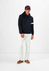 Mann iført en marineblå hettegenser med snorer, hvite bukser, brune slip-on sko og en mørkerød caps, stående mot en enkel hvit bakgrunn.