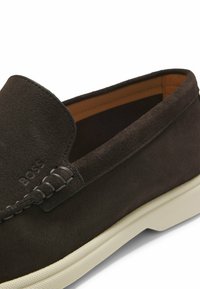 Gros plan sur une chaussure mocassin en daim marron foncé avec des détails cousus à la main visibles et une semelle en caoutchouc beige clair.