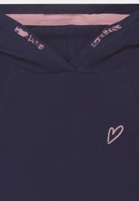 Námořnická modrá mikina s kapucí s výšivkou růžového srdce na hrudi, která má kontrastní růžovou podšívku a vzorované akcenty na stahovací šňůrce.