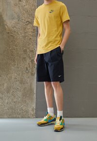 Yellow cotton t-shirt with green logo, black shorts with pockets, and multi-colored sneakers featuring blue and yellow accents.