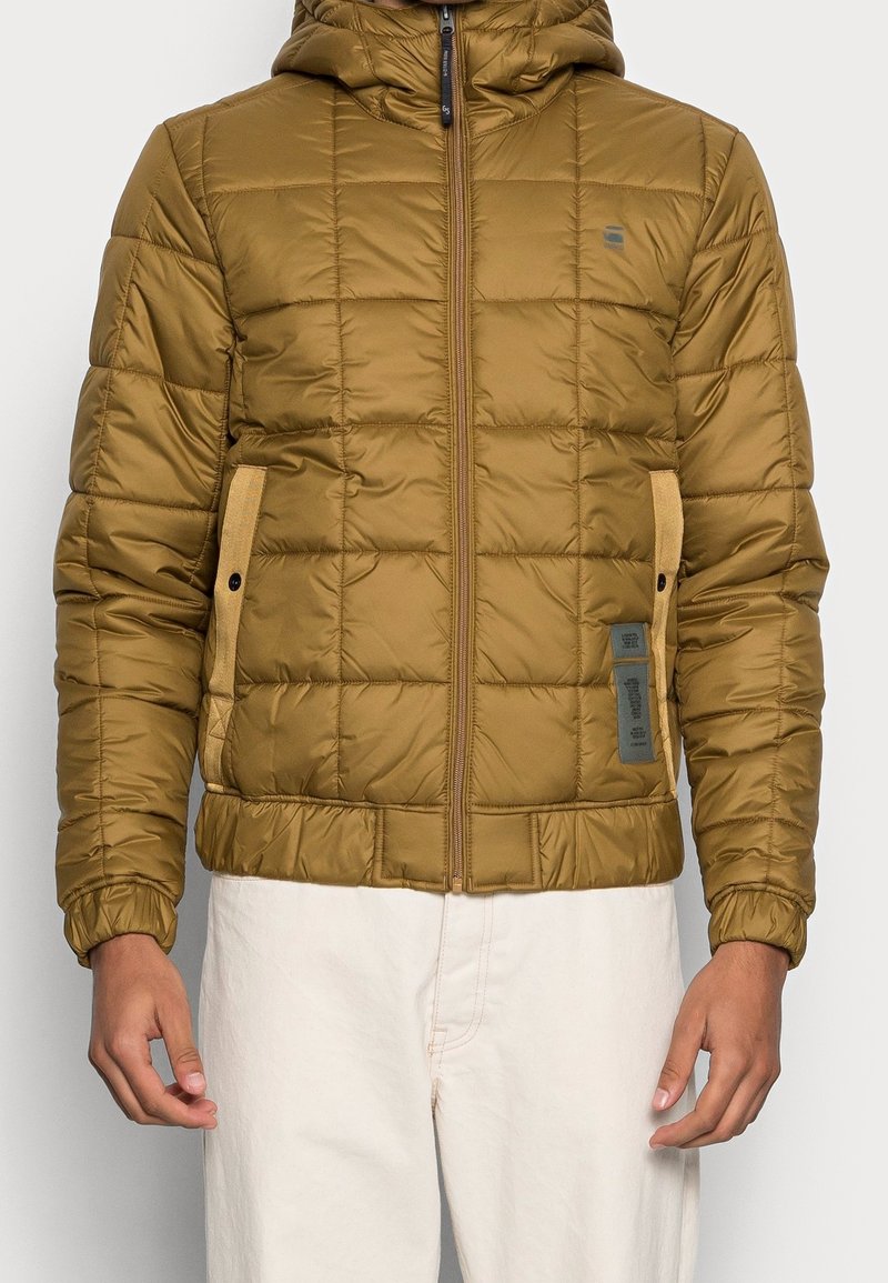 Overgångsjacka i olivgrönt, med dragkedja fram, huva, sidofickor och texturerat quiltat mönster. Detaljer inkluderar kontrasterande material och etiketter.