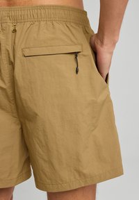 Calzoncini da bagno beige con vita elastica, tasca posteriore con zip e tessuto leggero e strutturato. I calzoncini hanno una vestibilità comoda e un taglio fino al ginocchio.