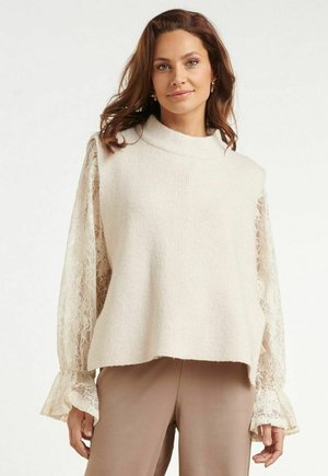 Vrouw met middellang bruin haar draagt een crèmekleurige mouwloze gebreide top over een kanten blouse met lange mouwen en beige broek, staand tegen een witte achtergrond.