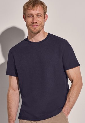 Homme souriant aux cheveux blonds courts portant une chemise à manches courtes bleu marine foncé et un pantalon brun clair, une main dans la poche.