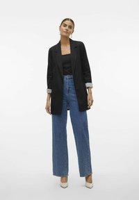 Blazer noir avec revers crantés, porté sur un haut noir. Jean bleu à taille haute avec jambes larges, délavé, associé à des talons hauts crème.