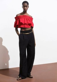 Top corto rosso scollato con strati arricciati, abbinato a pantaloni neri a vita alta con dettaglio di cintura in catena dorata.