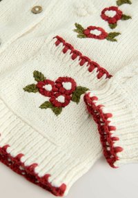 Sweter dziergany w kolorze kremowym z czerwonymi haftami kwiatowymi. Posiada ściągacze na rękawach i u dołu z dekoracyjnymi akcentami w czerwonym i białym kolorze.
