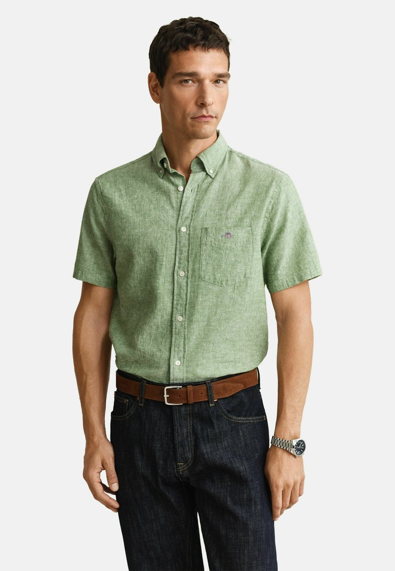 Mand iført en lysegrøn kortærmet skjorte med knapper, mørke jeans, brunt bælte og sølvfarvet håndledsur, stående foran en ensfarvet baggrund.